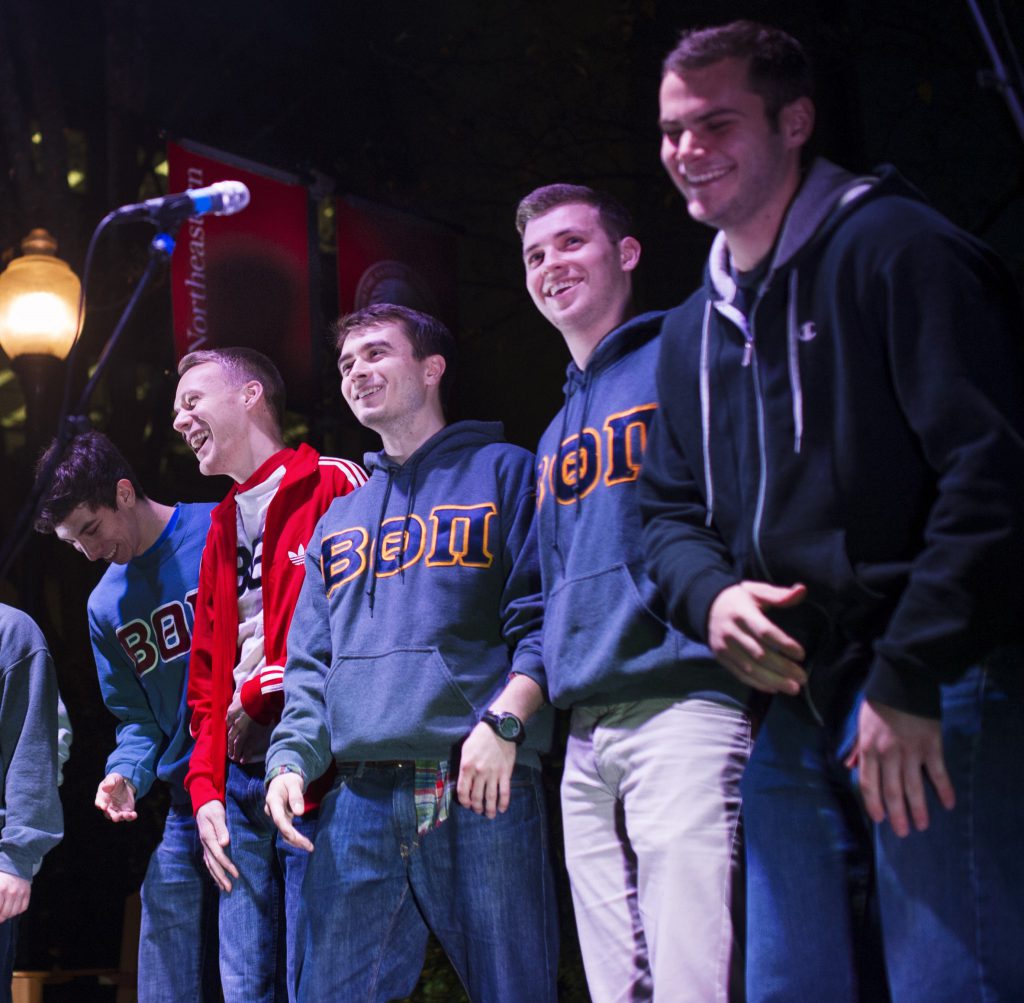 A group of 6 men in matching sweatshirts laughing and smiling. Their matching sweatshirts have the letters for their fraternity on them.