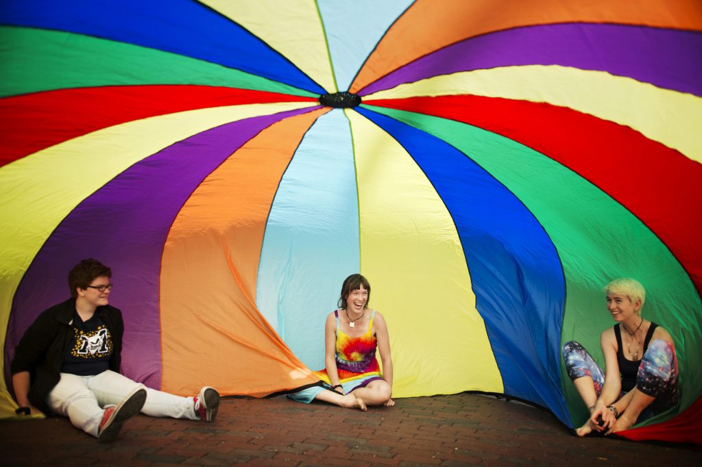 3 students sitting under a large rainbow parachute