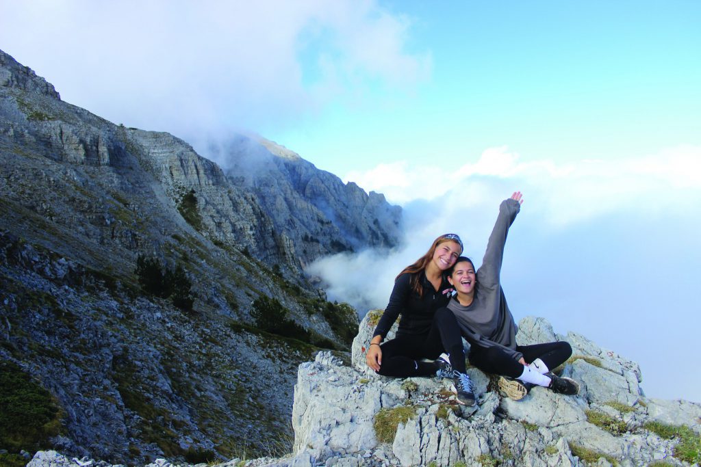 Two students sit on top of a large rock next to a mountain. They are smiling