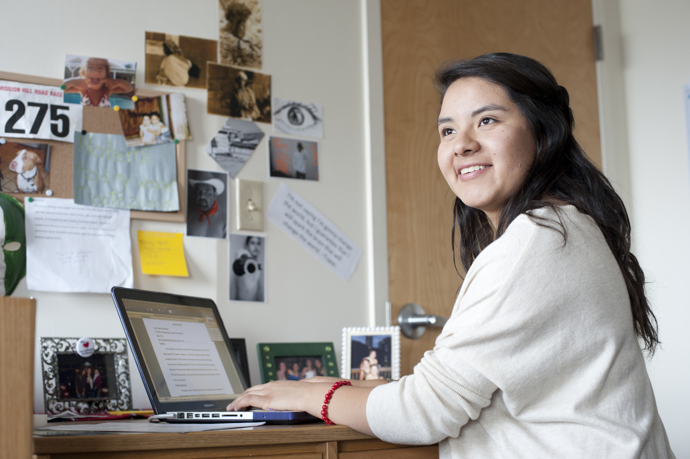 A female student in her dorm room, looking up and smiling with her laptop sitting on her desk.