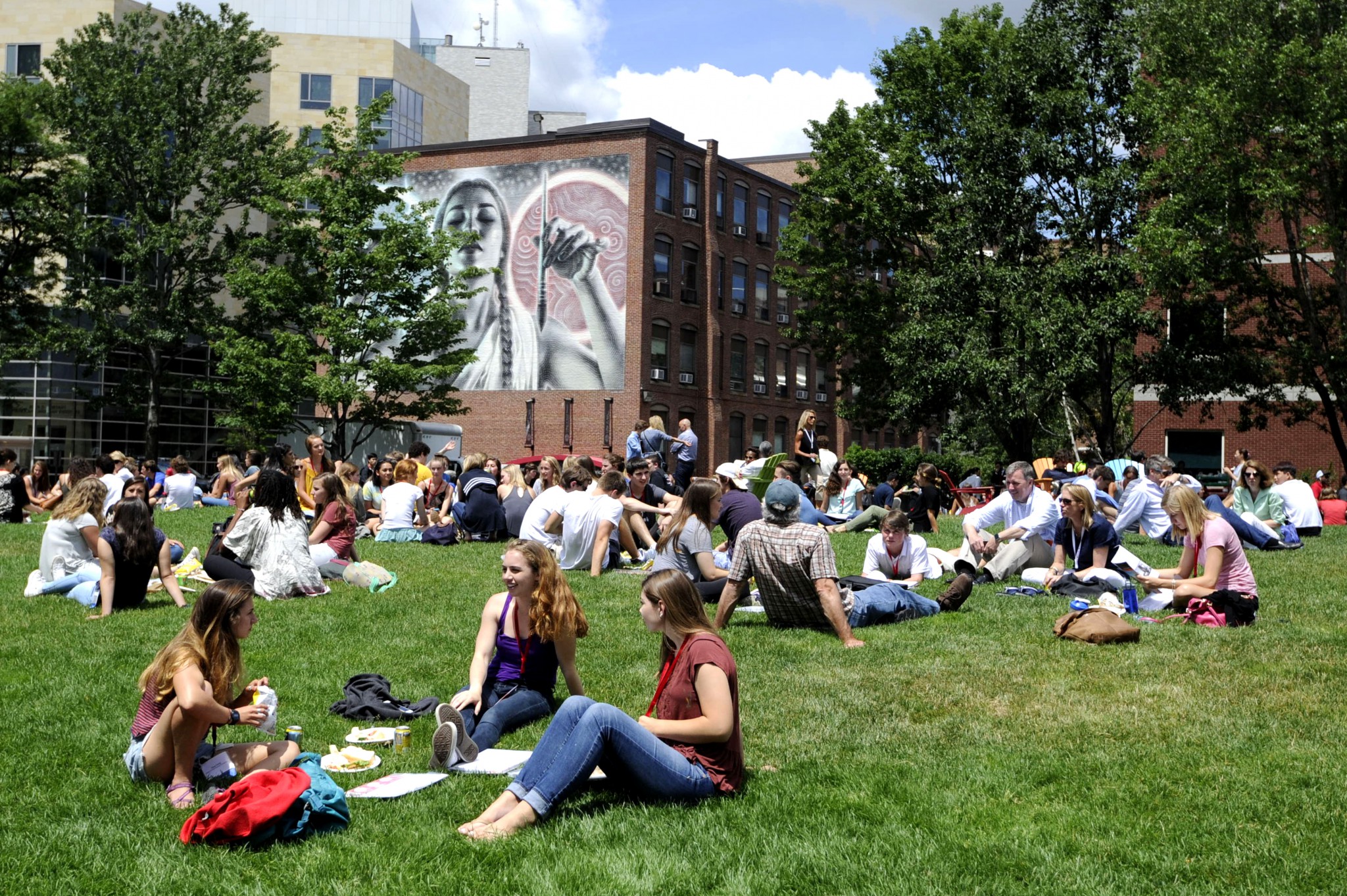Lots of students hanging out in a grassy area. There is a building in the background with a mural painted on the wall.