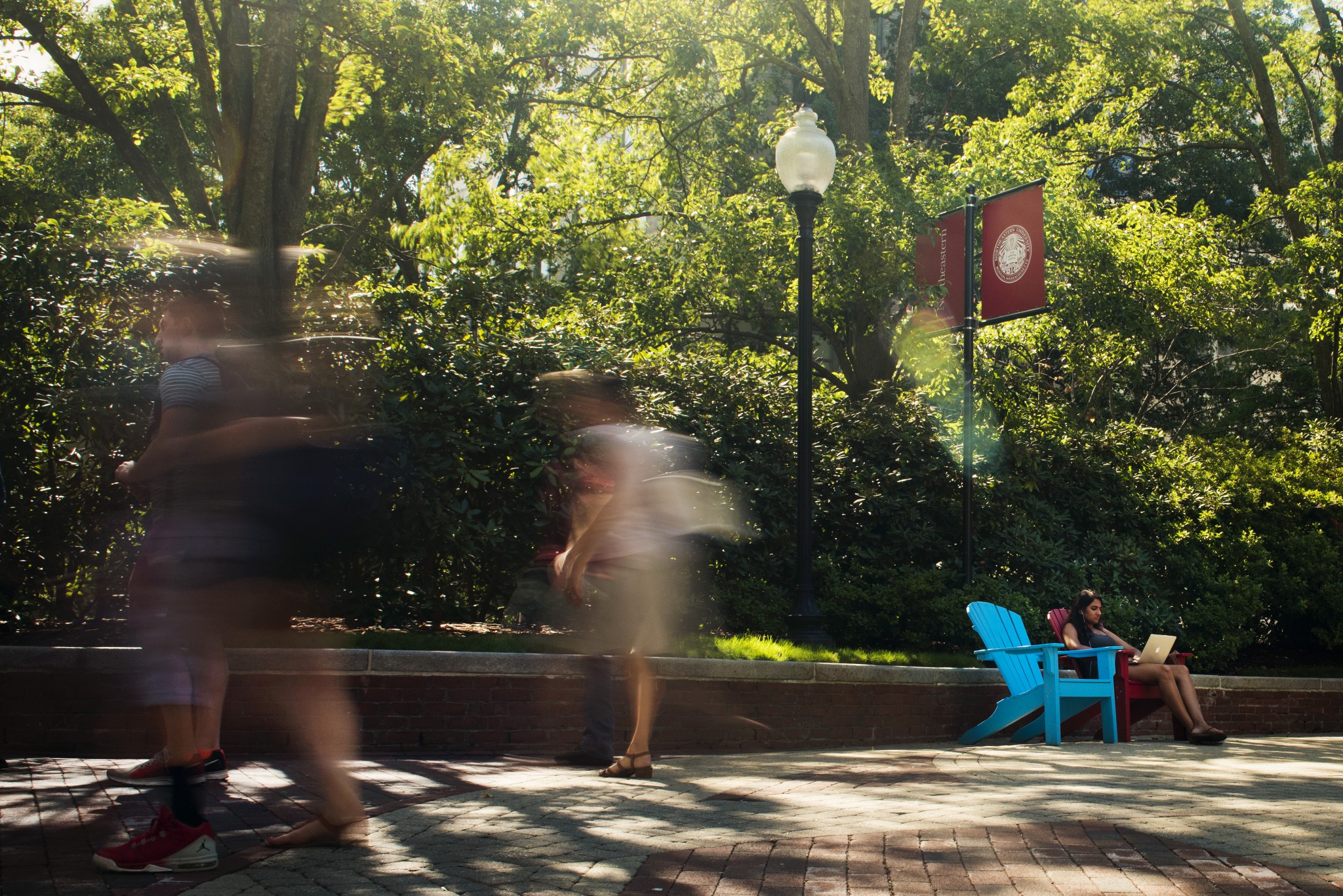 A blurry time lapse image of students on campus. There are blurry images of students walking in foreground, while the background showing a student sitting in a chair, trees and a Northeastern campus flag are clear.