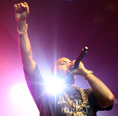 Picture of an entertainer speaking/singing into a microphone. He has one fist pumped in the air while bright lights shine behind him.